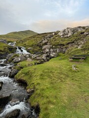 A serene landscape of Faroe Islands featuring a gentle stream flowing through lush green hills and rocky formations. The scene captures the peaceful beauty of nature under a partly cloudy blue sky.