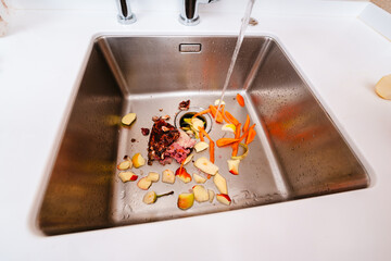 Food waste and vegetable scraps in a kitchen sink with running water and disposer.
