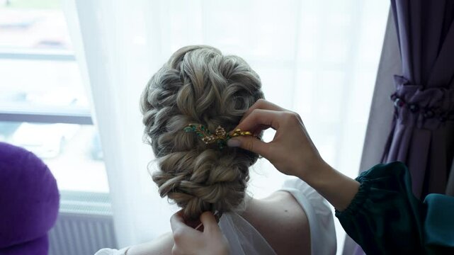 Panning of attaching the gold decorative bobby pin by female hand on head of woman in a dress with blond hair. Back view of nape and back, no face. Creating a wedding coiffure in the form of a bun