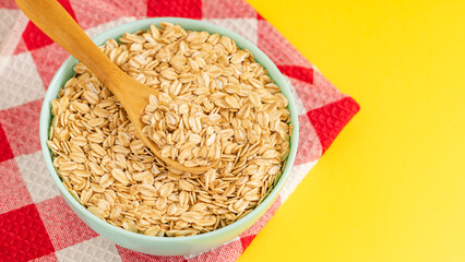 Pastel bowl filled with rolled oats and a wooden spoon on a red checkered cloth, set against a bright yellow background. Ideal for healthy food themes.