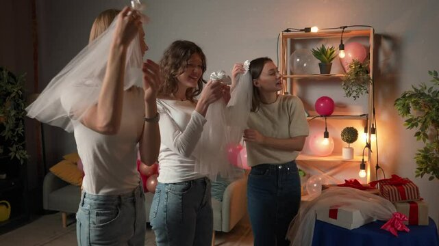 Group of young women friends and bride celebrating bachelorette party at home, waiting for bride, girls putting on the bride's veils in front of the mirror and talking.