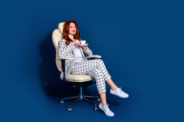 Stylish young woman with red hair relaxes in a checkered suit on a modern chair, sipping coffee...