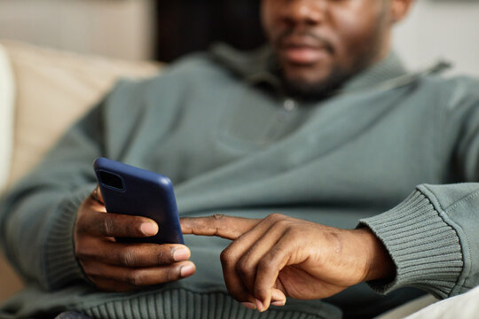 Close up of African American man holding phone in hand and tapping screen with finger while relaxing on couch at home, copy space