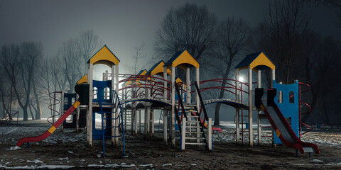 Empty children's playground at night, illuminated by soft streetlights, with colorful structures standing in contrast to the foggy and dark surroundings.