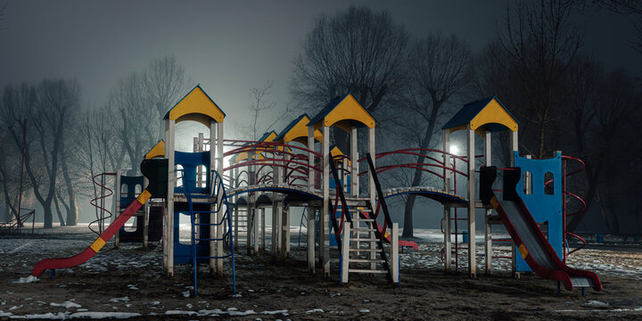 Empty children's playground at night, illuminated by soft streetlights, with colorful structures standing in contrast to the foggy and dark surroundings.