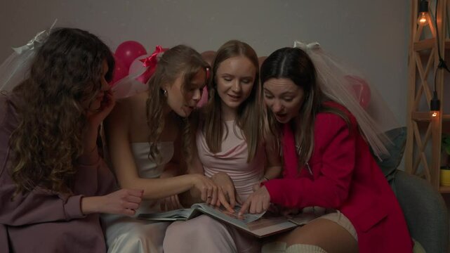 Group of young women friends and bride celebrating bachelorette party at home, bride and bridesmaids sit on sofa and look through old photo albums, happy memories.