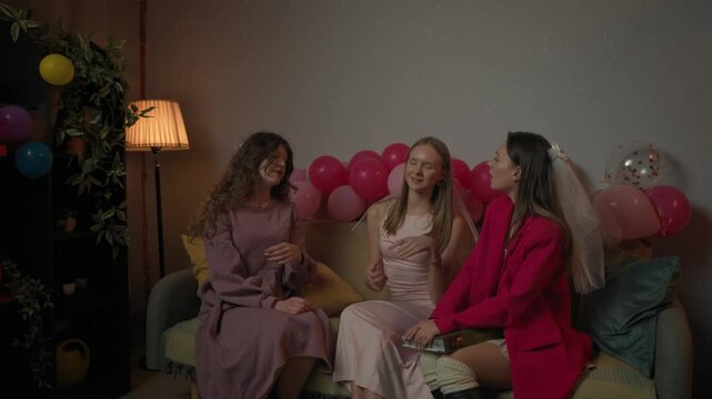 Group of young women friends and bride celebrating bachelorette party at home, bridesmaids sit on sofa holding photo album and talk, waiting for bride.