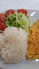 Close up do prato branco com omelete, arroz branco, batata frita, salada verde, e tomate.