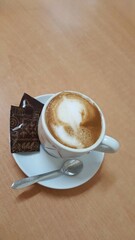 Uma xícara branca de capuccino com dois pacotinhos pretos de açúcar, pires branco e colher, sobre mesa marrom.