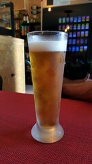 Copo de cerveja, fino ou imperial, de portugal, sobre mesa vermelha, e imagens desfocadas ao fundo, do interior de um bar.