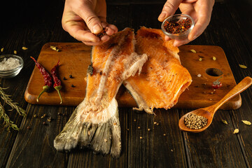 Hands are carefully sprinkling spices over a freshly sliced fish fillet on a wooden cutting board....