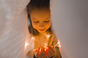 A cute little girl in a knitted sweater is hiding under a white blanket with a magical Christmas light. A cozy Christmas and New Year. Home comfort and joy, miracle