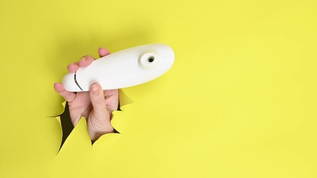 Female hand holding white clitoral vacuum vibrator sticking out of yellow paper background. 