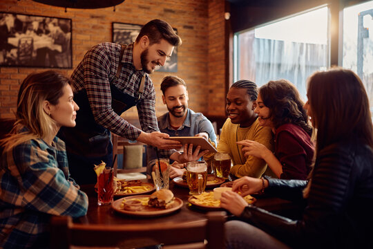 Happy waiter using touchpad while showing menu to group of guests in pub.