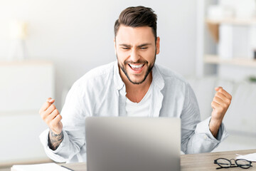 Portrait of overjoyed guy celebrating success, shaking clenched fists while sitting at desk in living room interior, copy space. Man using laptop computer, reading great news, screaming yes