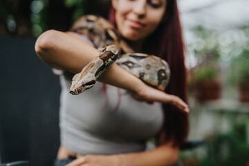 Fototapeta premium A girl with long hair smiles gently as a boa constrictor comfortably rests on her arm, surrounded by lush greenery.