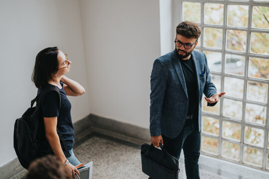 A professor and student engage in conversation, standing near a large window in a bright corridor. The scene captures an educational setting, emphasizing mentorship and learning in a casual