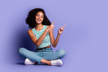 Charming young woman in casual outfit sitting cross-legged on purple backdrop with playful pointing...