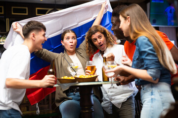 Multiracial Russia sports fans, men and women, supporting their favourite team in bar, raising state flag and screaming chants together.