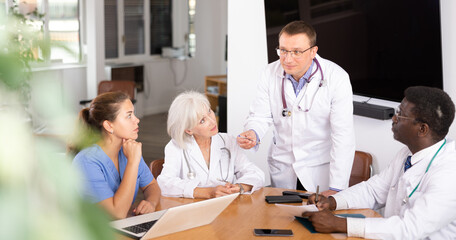 Group of different people medics in medical uniforms having discussion about work at table in office