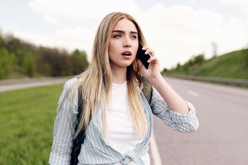 Confused young blonde woman standing along road, feeling lost, calling on cellphone, asking for...
