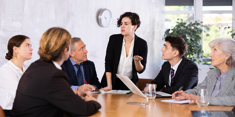 Young Latin female turning on presentation on projector while office coworkers of different ages sitting at table with laptops and notes