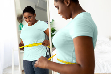 Plus size millennial black woman measuring waist with tape, standing in front of mirror, satisfied with results of slimming diet at home. Young African American lady achieving success in weight loss