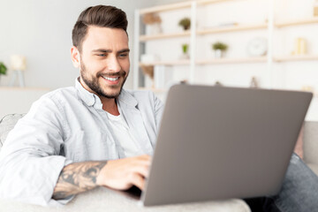 Modern job lifestyle. Happy tattooed man sitting on sofa at home and working on laptop computer...