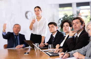 Office coworkers of different ages watching presentation while sitting at table with laptop and notes
