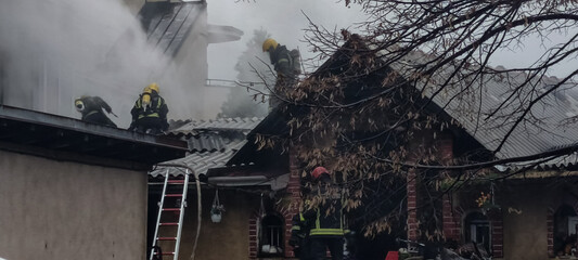 Firefighters climbing a ladder to the roof of a house, smoke coming out from behind and around it