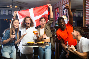 Obraz premium Group of Denmark football team fans spending time in bar, drinking bear and having fun. People with state flag in pub.