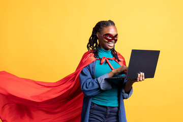 Smiling african american girl portraying superhero hiding identity, working on laptop, studio...