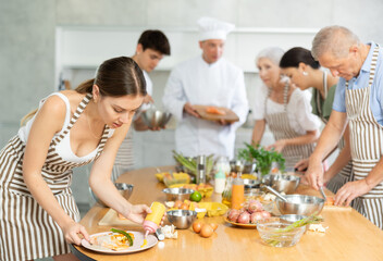 Positive young woman in striped apron participating in group cooking class, standing at table and seasoning prepared dish with sauce ..