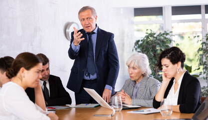 Annoyed senior male boss yelling at office colleagues of different ages taking notes at table with laptops