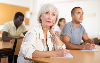 Positive interested elderly woman listening attentively and taking notes on lesson during language...