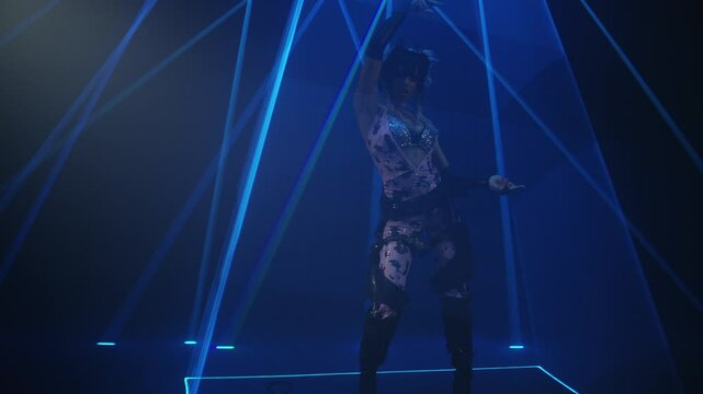Woman wearing unconventional clothing stands on a dark stage with laser lights moving during a music video