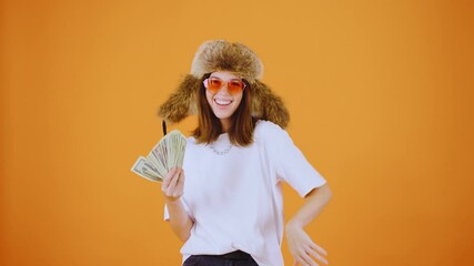 A stylish woman confidently exudes charisma as she poses with cash and trendy sunglasses and in a stylish fur hat against a vibrant orange background