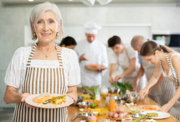 Happy smiling senior woman participating in group cooking class, standing in striped apron with plate of prepared dish in hands..