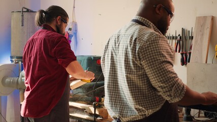 Team of woodworkers in assembly shop using sandpaper and spindle moulder for sanding wooden surface before painting it. Carpenters wearing safety glasses using abrasive sponges on wood, camera A