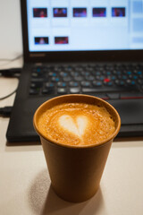 Cappuccino in brown paper disposable cup on table near laptop. Soft selective focus