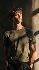Obraz premium A male model poses in an olive green t-shirt, standing against a textured wall with gentle sunlight streaming through. The atmosphere is serene, highlighting contemporary style and mood