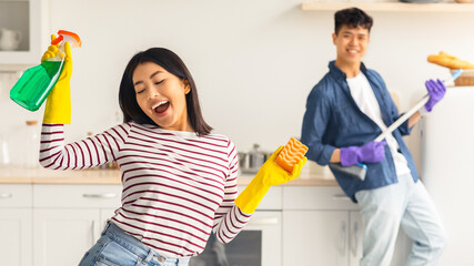 Funny asian woman singing songs and dancing with closed eyes while cleaning kitchen at home,...