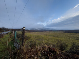 Rural Landscape with Weathered Fence, Open Field, and Distant Mountain Covered in Mist Under Cloudy...