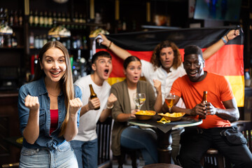 Cheerful international sport football fans waving German flag and drinking beer, eating chips in the sport bar