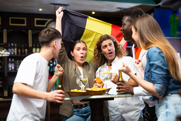 Group of emotional young adults friends, football fans cheering for favorite Belgian team together while watching match in sports bar..