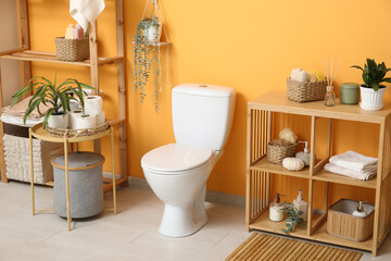 Interior of stylish restroom with white toilet bowl, coffee table and shelving unit near orange wall