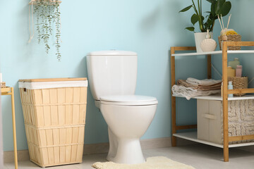 Interior of stylish restroom with ceramic toilet bowl, houseplant, braided basket and shelving unit near blue wall