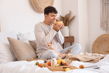 Young man with cup of tea and cute cat in bedroom on autumn day