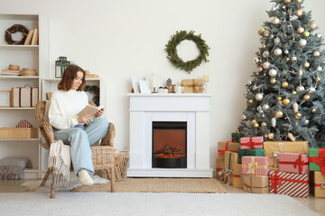 Young woman reading book in armchair near fireplace at home on Christmas eve