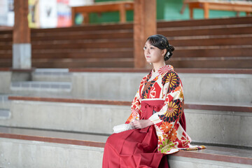 Japanese woman in her 20s wearing red hakama, staple for Japanese university graduates, sits in audience seats in large room.